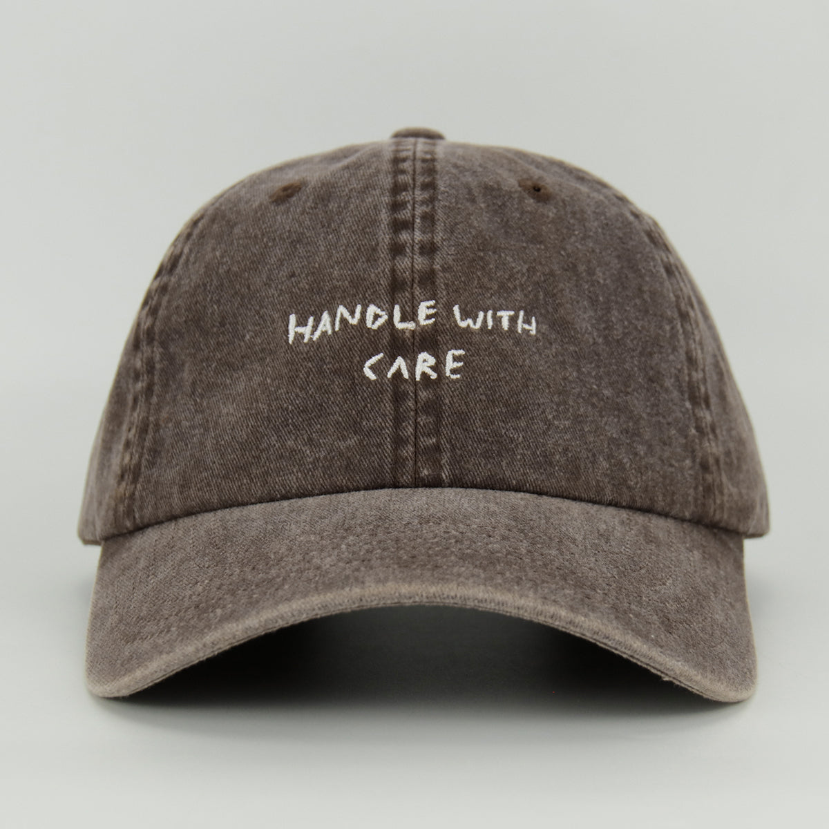 A brown cotton cap with the phrase 'HANDLE WITH CARE' embroidered in white on the front.