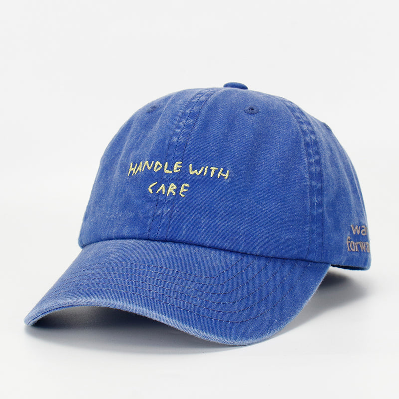 A blue cotton cap with the phrase 'HANDLE WITH CARE' embroidered on the front.