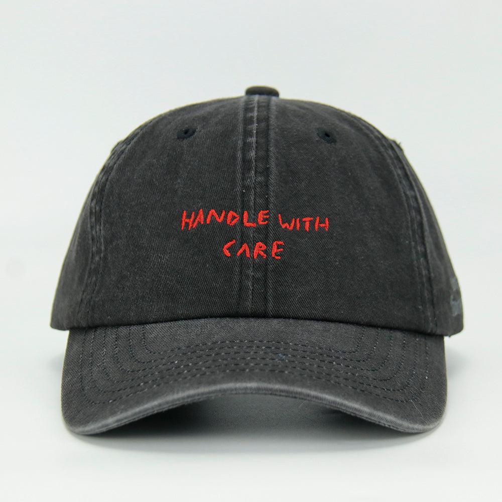 A black cotton cap with the phrase 'HANDLE WITH CARE' embroidered in red on the front.