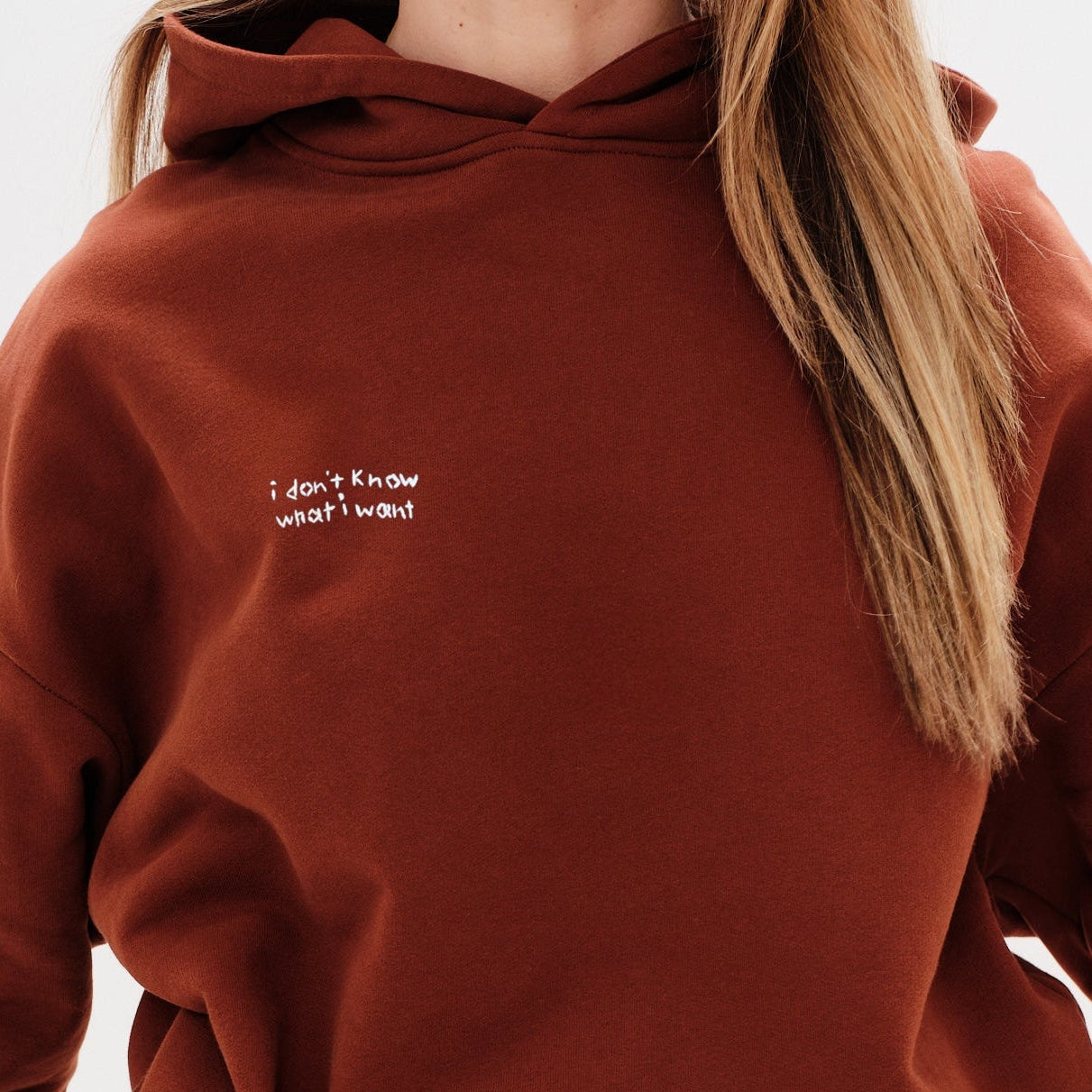 Person wearing a brown hoodie with a small white text I don't know what I want embroidery