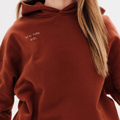 Woman wearing a brown hoodie with a New York Girl embroidery.